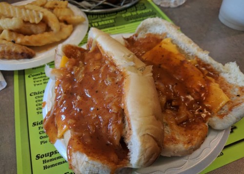 Two hot dog buns filled with chili and cheese, served with a side of crinkle-cut fries on a colorful menu background.
