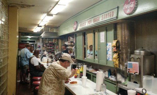 A bustling diner kitchen with patrons at the counter, vintage decor, and a menu sign overhead.