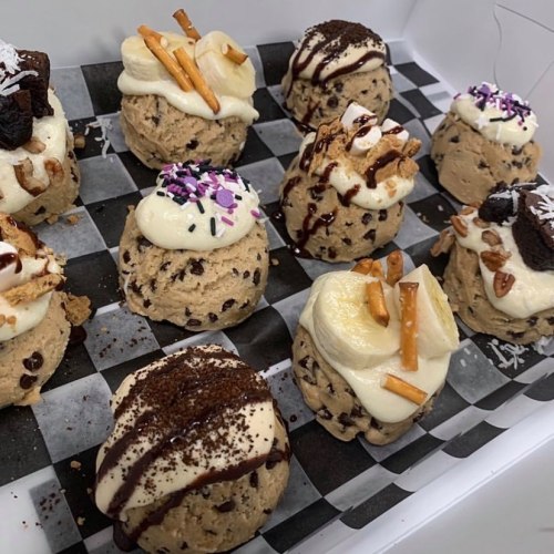 A variety of cookie dough desserts topped with frosting, pretzels, chocolate, and sprinkles on a checkered paper.