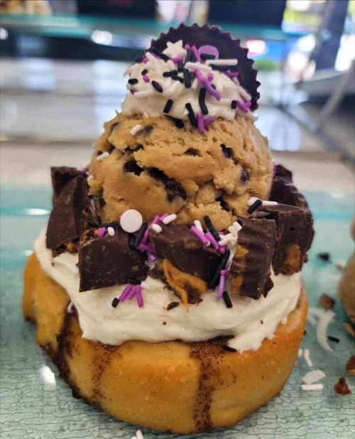 A dessert featuring a cookie dough scoop on a cupcake, topped with whipped cream, chocolate pieces, and colorful sprinkles.