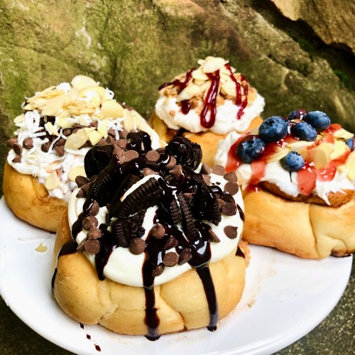 A plate of four decadent cinnamon rolls topped with cream, chocolate chips, fruit, and nuts against a natural backdrop.