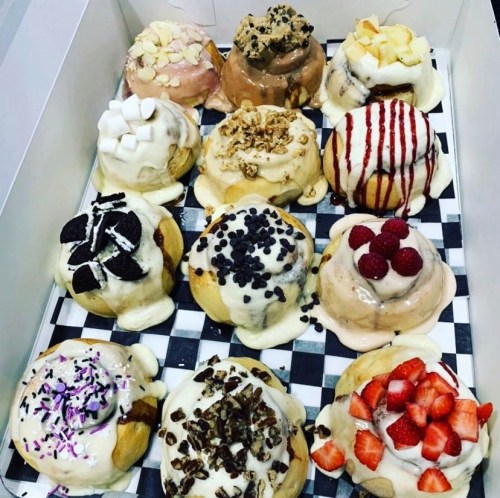 A box of twelve colorful cinnamon rolls topped with various fruits, nuts, and drizzles, arranged on a checkered paper.