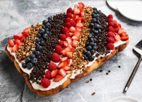 A rectangular dessert topped with layers of strawberries, blueberries, raspberries, chocolate chips, and granola on cream.