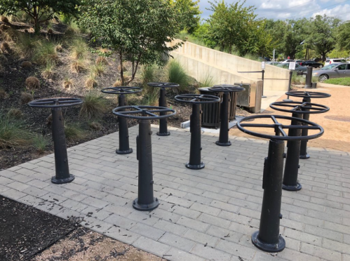 A row of black metal bike racks on a paved area surrounded by grass and trees.