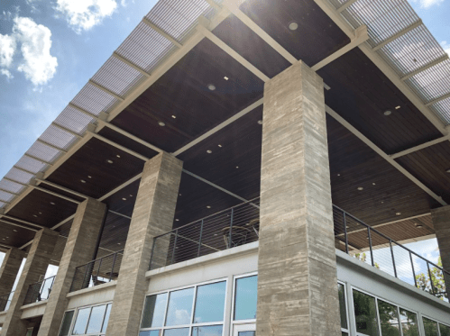 Modern building exterior with large columns, a wooden ceiling, and a balcony under a bright blue sky.