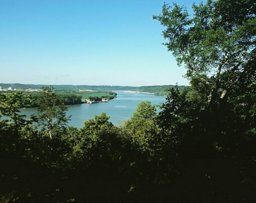 A serene river view surrounded by lush greenery and distant hills under a clear blue sky.