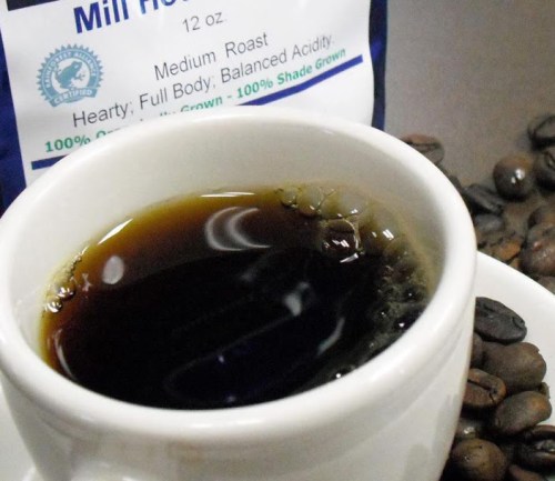 A close-up of a white cup filled with dark coffee, surrounded by coffee beans and a bag of medium roast coffee.