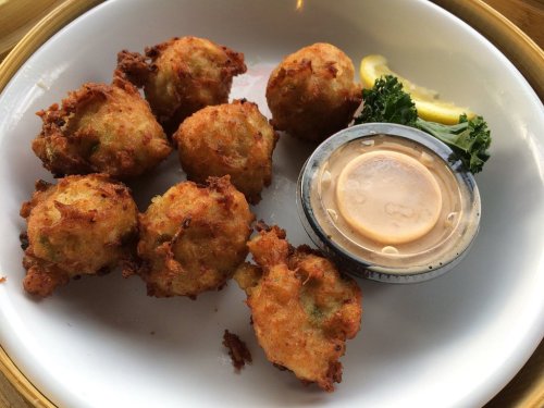 A plate of golden-brown fritters served with a dipping sauce and a lemon wedge, garnished with a piece of kale.