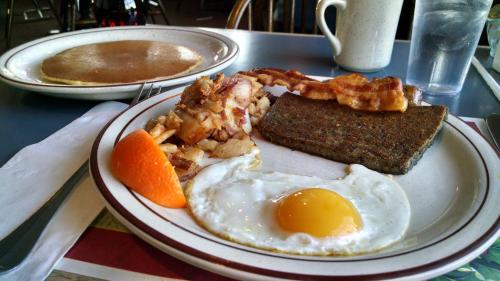 A plate of breakfast featuring an egg, bacon, hash browns, and an orange slice, with a pancake and coffee in the background.