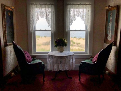 A cozy room with two chairs and a table, overlooking a beach through lace curtains. Flowers adorn the table.