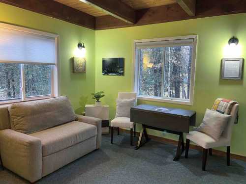 Cozy room with a green wall, a sofa, a desk, two chairs, and large windows overlooking trees.