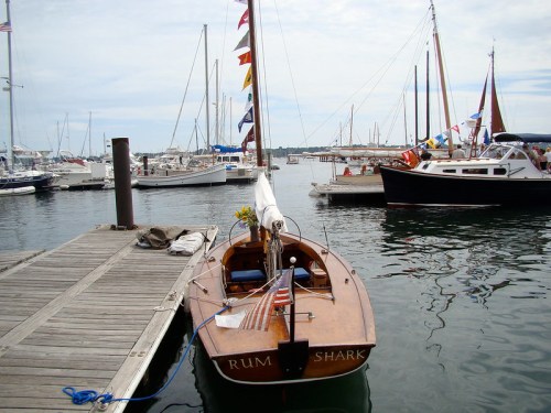 A small wooden boat named "RUM SHARK" docked at a marina, surrounded by larger sailboats and a cloudy sky.
