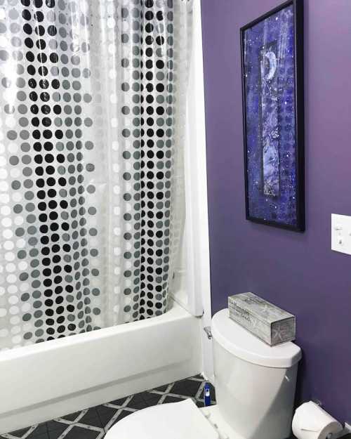 A bathroom with a purple wall, a polka dot shower curtain, and a decorative framed piece above the toilet.