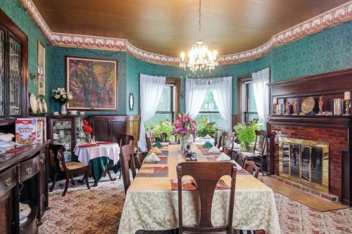 Elegant dining room with a large table, green walls, floral decor, and a cozy fireplace. Bright natural light filters in.
