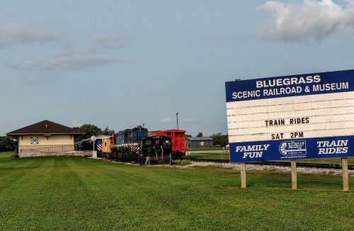 A train parked beside a museum sign advertising scenic train rides, set in a grassy area under a cloudy sky.