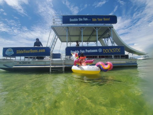 A colorful pontoon boat with a water slide, featuring a child on a float in the water, under a bright blue sky.