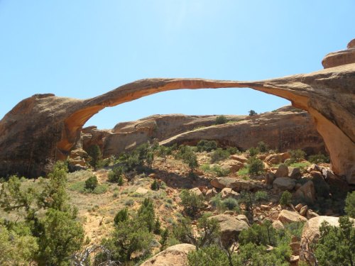Natural rock arch spanning a desert landscape, surrounded by rugged terrain and sparse vegetation under a clear blue sky.