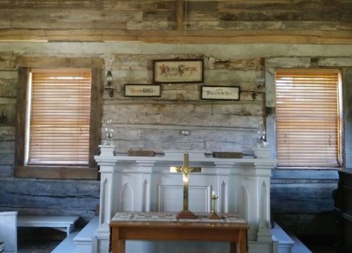 Interior of a rustic chapel with wooden walls, a white altar, and two windows with wooden blinds.