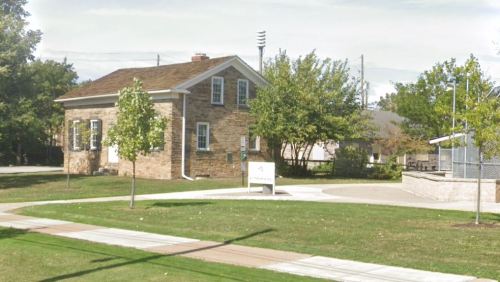 A stone building surrounded by trees and grass, with a pathway leading to a nearby structure and sign.