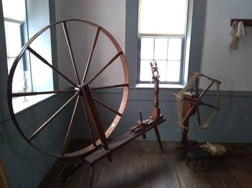 Two traditional spinning wheels in a well-lit room with wooden floors and windows.