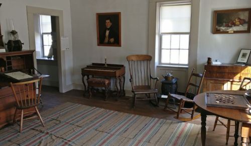 A cozy room with wooden furniture, a rug, and a painting on the wall, featuring a desk, chairs, and a small table.