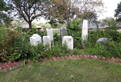 A cluster of old gravestones surrounded by overgrown plants and grass in a park-like setting.