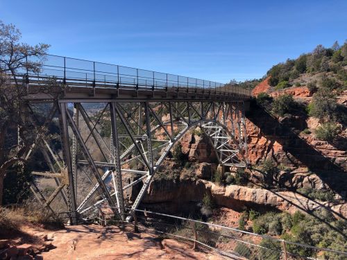 A metal bridge spans a rocky canyon, surrounded by trees and blue sky, showcasing a scenic landscape.