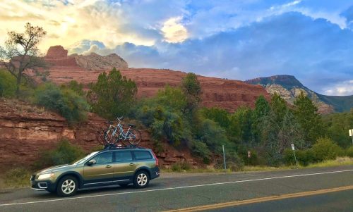 A car parked by the roadside with bicycles on the roof, set against a backdrop of red rock formations and a cloudy sky.