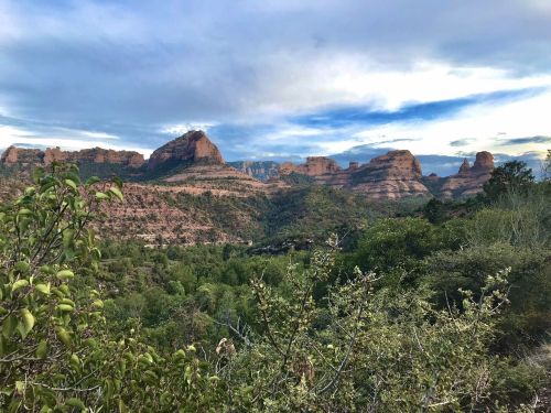 Scenic view of red rock formations and lush greenery under a cloudy sky in a mountainous landscape.