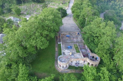 Aerial view of an abandoned building surrounded by trees, near a cemetery and a winding road.
