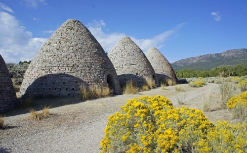 Stone kilns with conical roofs stand along a gravel path, surrounded by yellow wildflowers and mountains in the background.