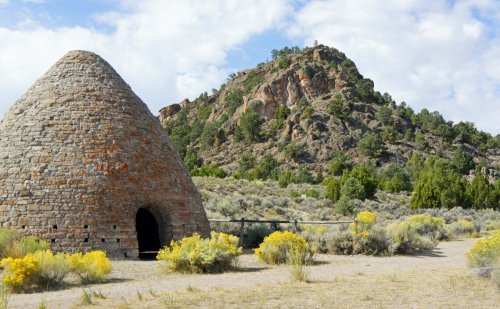 A stone beehive-shaped kiln stands in a desert landscape with a rocky hill and scattered yellow flowers in the foreground.