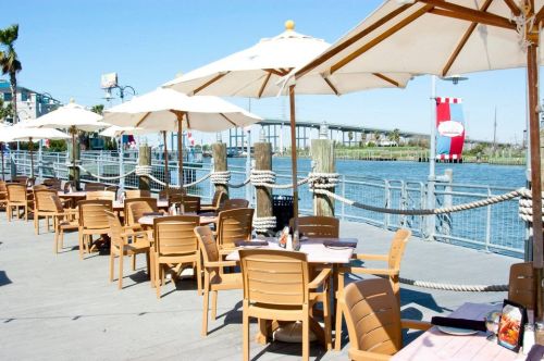 Outdoor dining area with wooden tables and umbrellas by a waterfront, featuring a clear blue sky and distant bridge.