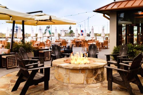 A cozy outdoor seating area with a stone fire pit surrounded by wooden chairs, overlooking a waterfront dining space.