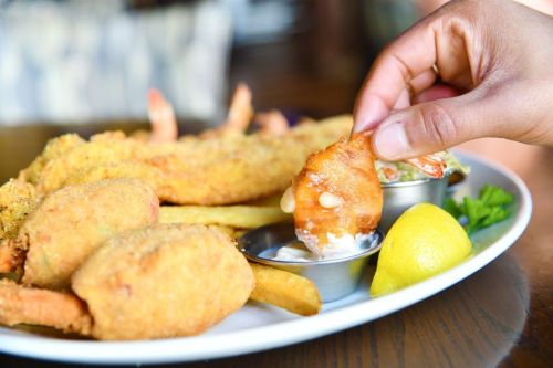 A hand dips a fried shrimp into a small bowl of sauce, surrounded by fried seafood, fries, and a lemon wedge.