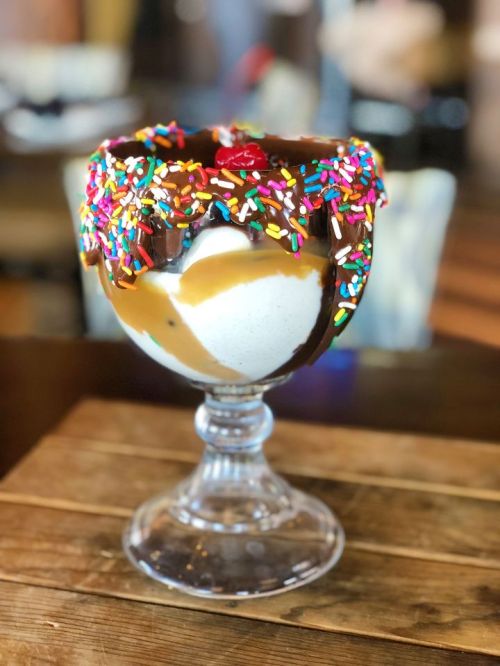 A tall glass dessert filled with ice cream, topped with chocolate, caramel, sprinkles, and a cherry.