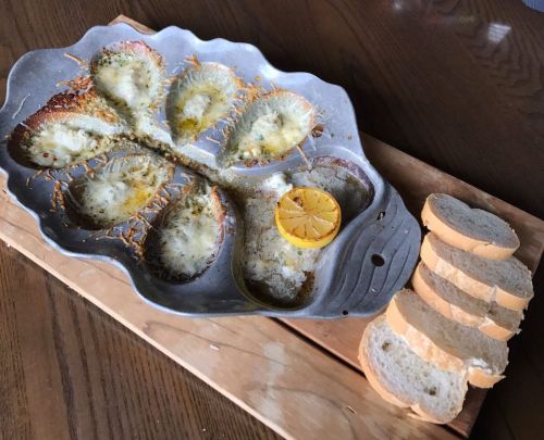A silver dish with baked cheese and a lemon slice, accompanied by slices of bread on a wooden board.