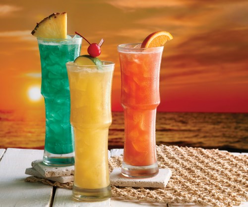 Three colorful cocktails with fruit garnishes, set against a sunset over the ocean.