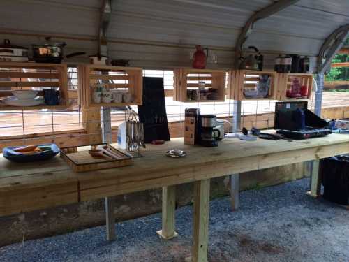 A rustic outdoor kitchen with wooden shelves, pots, and utensils, featuring a long table and a chalkboard sign.
