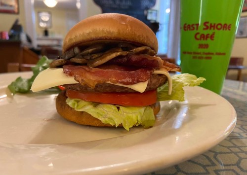 A delicious burger with lettuce, tomato, bacon, cheese, and grilled onions on a bun, served with a green drink.
