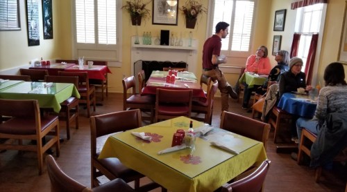 A cozy restaurant interior with colorful tables, a waiter serving, and patrons enjoying their meals.