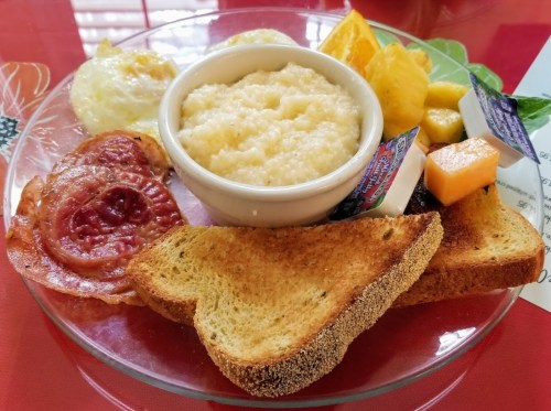 A plate with scrambled eggs, grits, bacon, toast, and fruit, served with butter and jam.