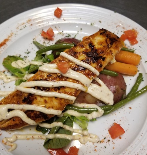 Grilled fish fillet on a bed of red potatoes, green beans, and carrots, drizzled with sauce and garnished with tomatoes.