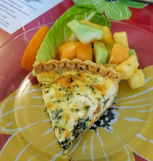 A slice of quiche with spinach and cheese, served with a side of mixed fruit including melon and pineapple.