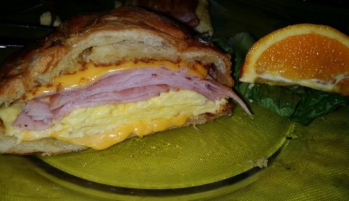 A close-up of a croissant sandwich filled with ham and melted cheese, served with a slice of orange and lettuce.
