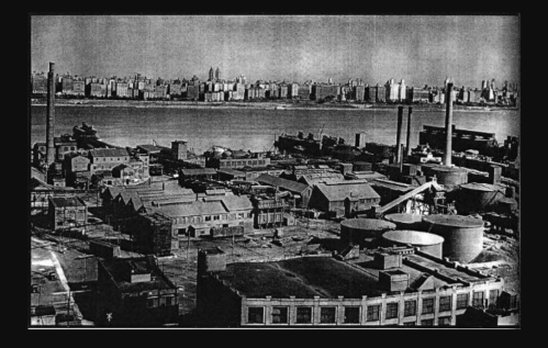 Black and white aerial view of an industrial area with factories and a city skyline in the background.