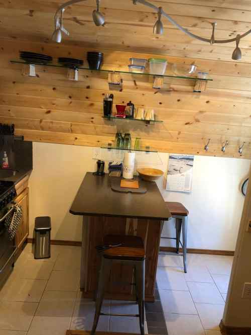 A cozy kitchen with wooden walls, a central island, shelves of dishes, and modern lighting.