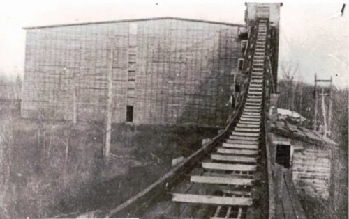 A vintage black-and-white photo of a wooden roller coaster track leading to a large, partially constructed building.
