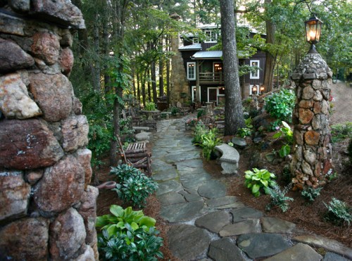 A stone pathway leads to a charming house surrounded by lush greenery and trees, with lanterns illuminating the path.