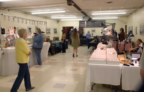 A spacious indoor market with vendors displaying jewelry and crafts, and visitors browsing the stalls.